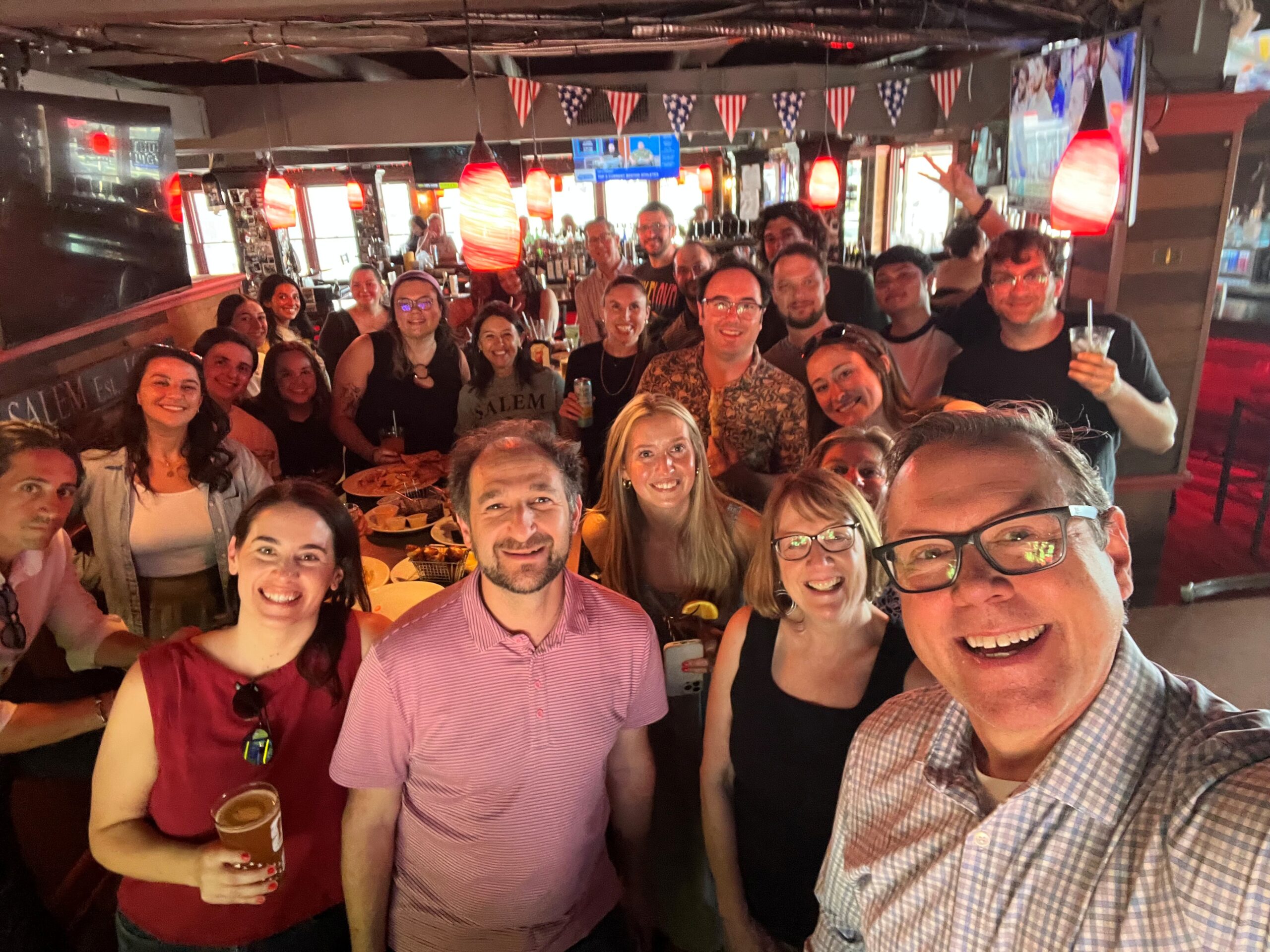 A candid group selfie of the combined Arketi and Sperling team gathered inside a lively restaurant, with team members smiling around tables and standing together in an informal setting.